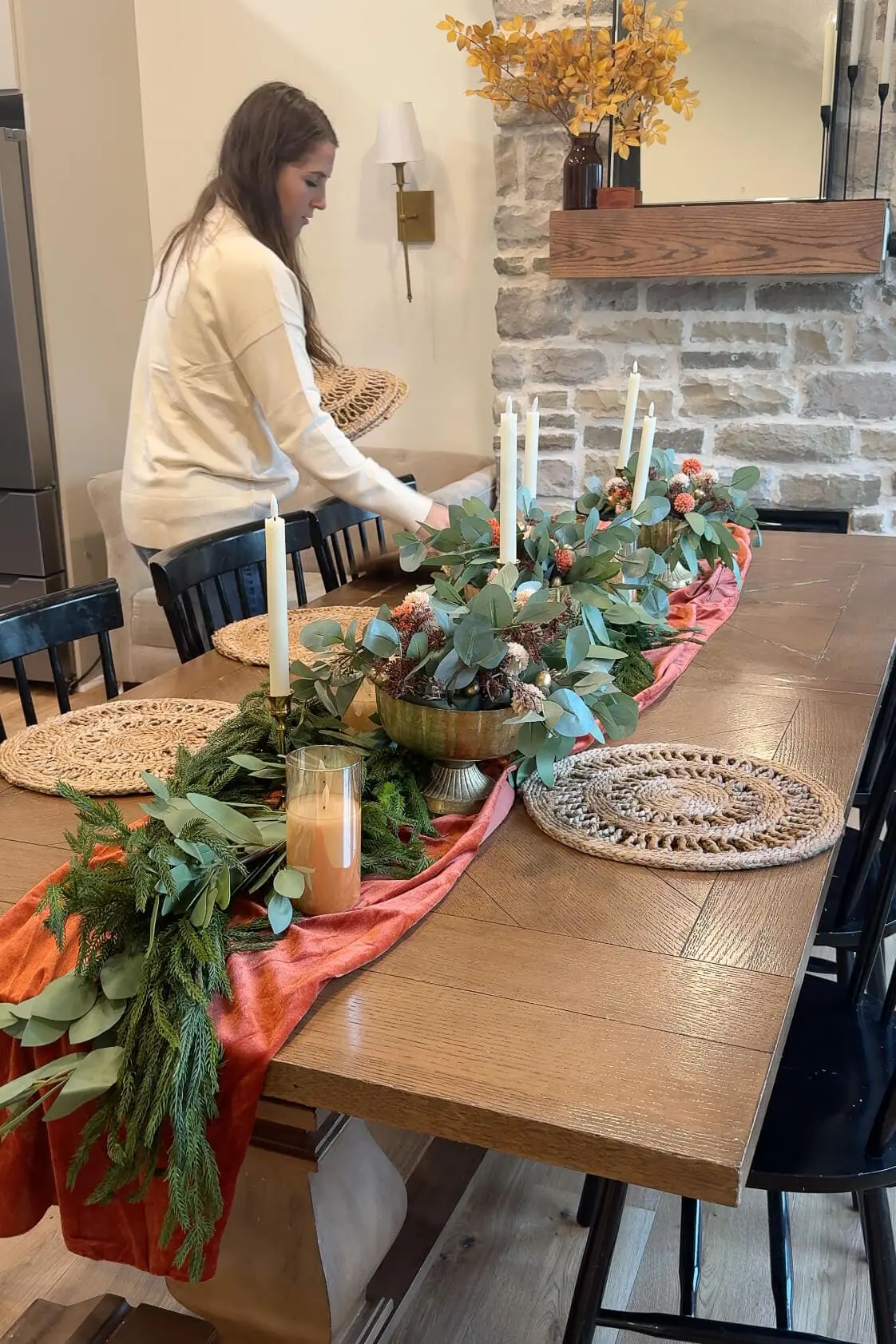 Woman adding round woven placemats to a dining table decorated with a festive tablescape featuring greenery, candles, and orange accents.